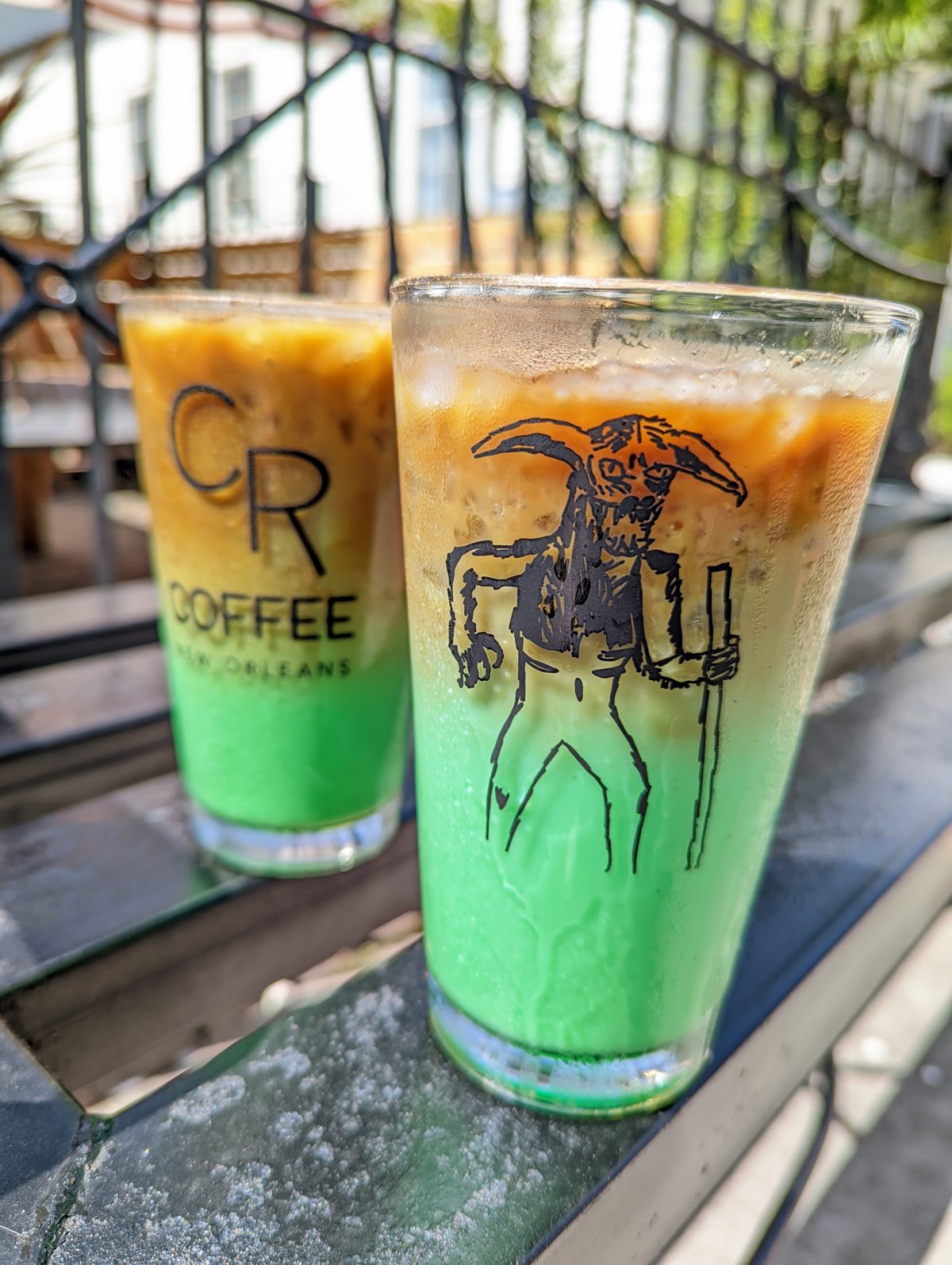 Two Bayou Beast iced lattes side by side on the patio rail at CR Coffee, layered coffee brown over bright pandan green, the front glass printed with a hand drawn horned beast and the glass behind printed with the CR Coffee logo