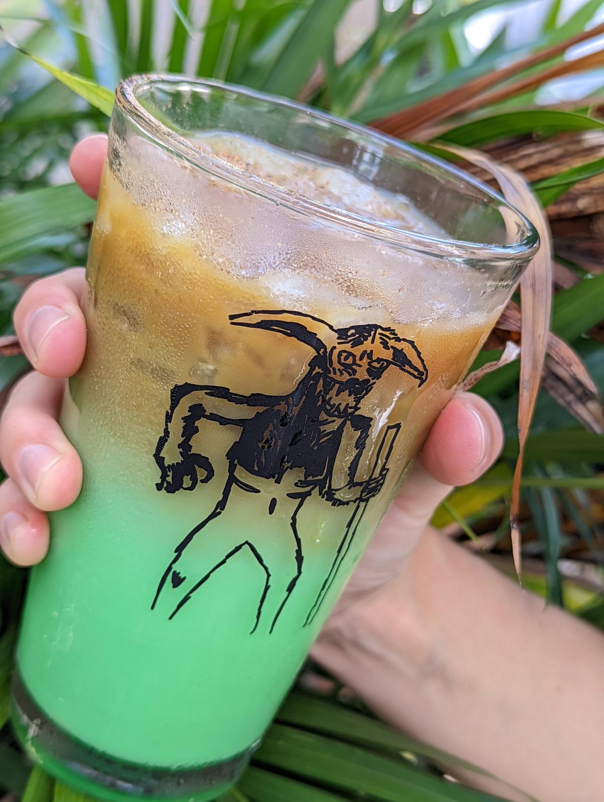 A hand holding a Bayou Beast iced latte in front of palm fronds, the drink shading from coffee brown at the top to bright green at the bottom, with a hand drawn horned beast on the glass