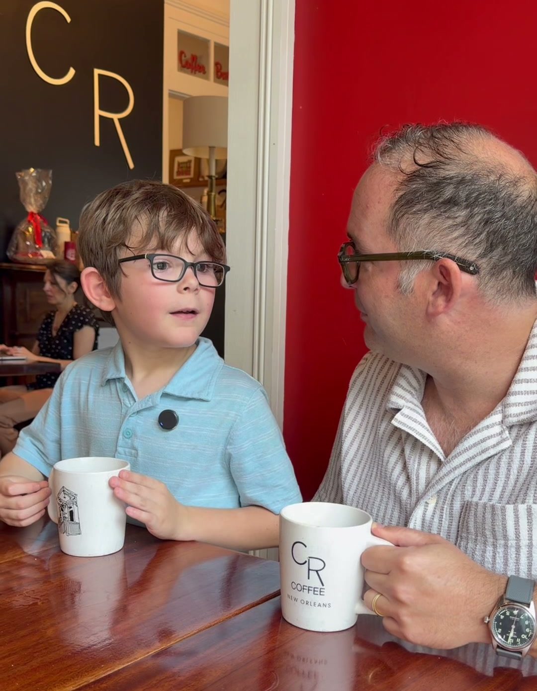 Kevin Pedeaux and his seven-year-old son Philip sitting at a wooden table at CR Coffee Shop, each with a white mug, in front of the black CR logo wall and red wall