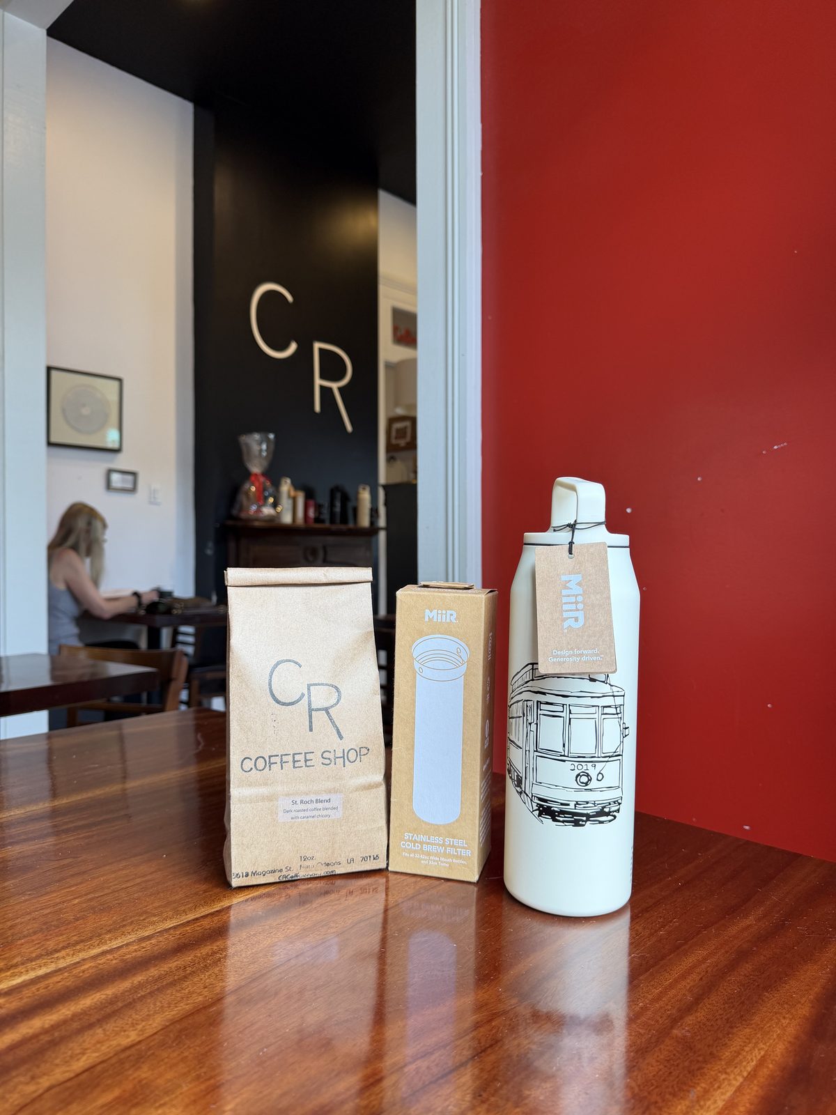 The CR Coffee Cold Brew Kit on a wood table: a bag of St. Roch Blend coffee, a boxed stainless steel cold brew filter, and a white MiiR insulated bottle printed with a New Orleans streetcar, with the red wall and CR monogram of the shop behind them