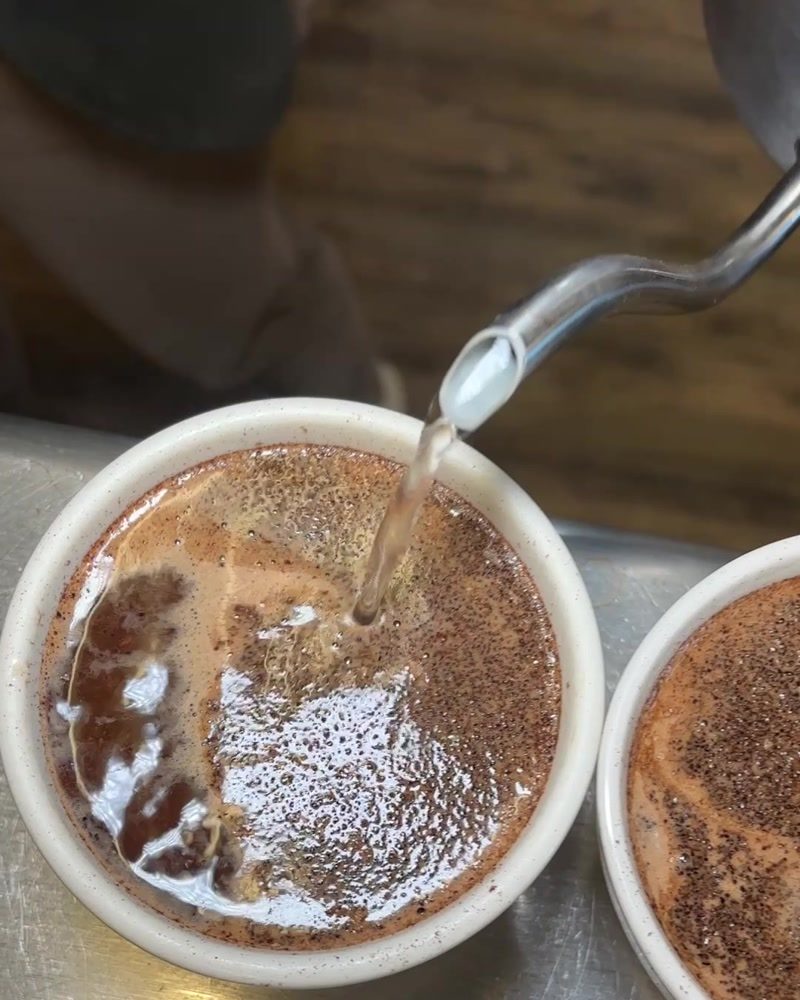 Hot water pours from a gooseneck kettle into a ceramic cupping bowl, the ground coffee blooming and forming a crust on the surface