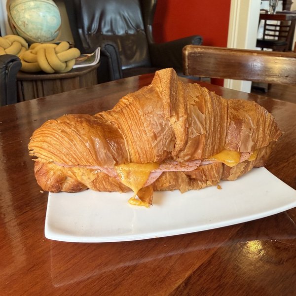 A ham and cheese croissant on a white plate, cheese melted through and running out the side onto the plate, on a polished wood table in the shop