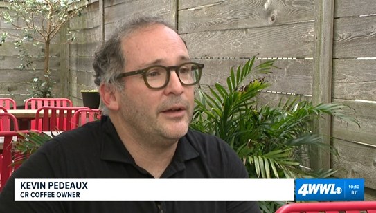 Kevin Pedeaux in glasses and a black polo, seated on the patio at CR Coffee Shop among red chairs and greenery, with a WWL Louisiana lower-third reading Kevin Pedeaux, CR Coffee Owner